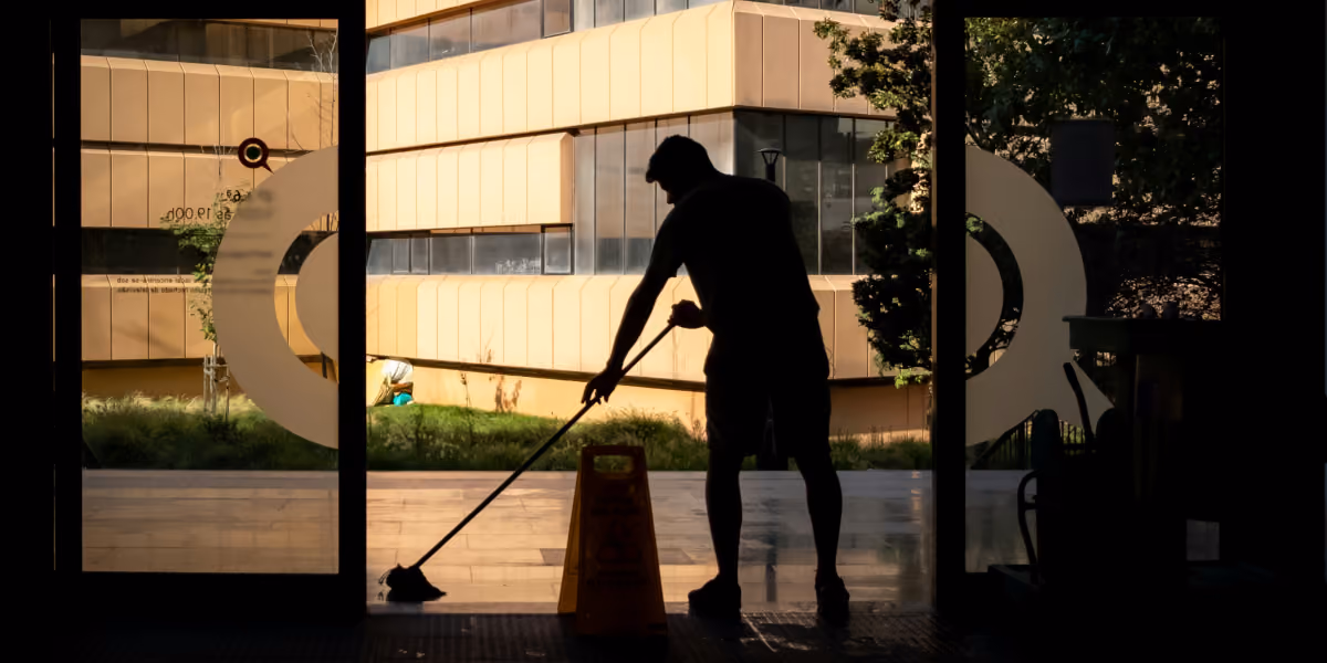 Silhouette of a person mopping a floor near glass doors with a yellow caution sign outside.