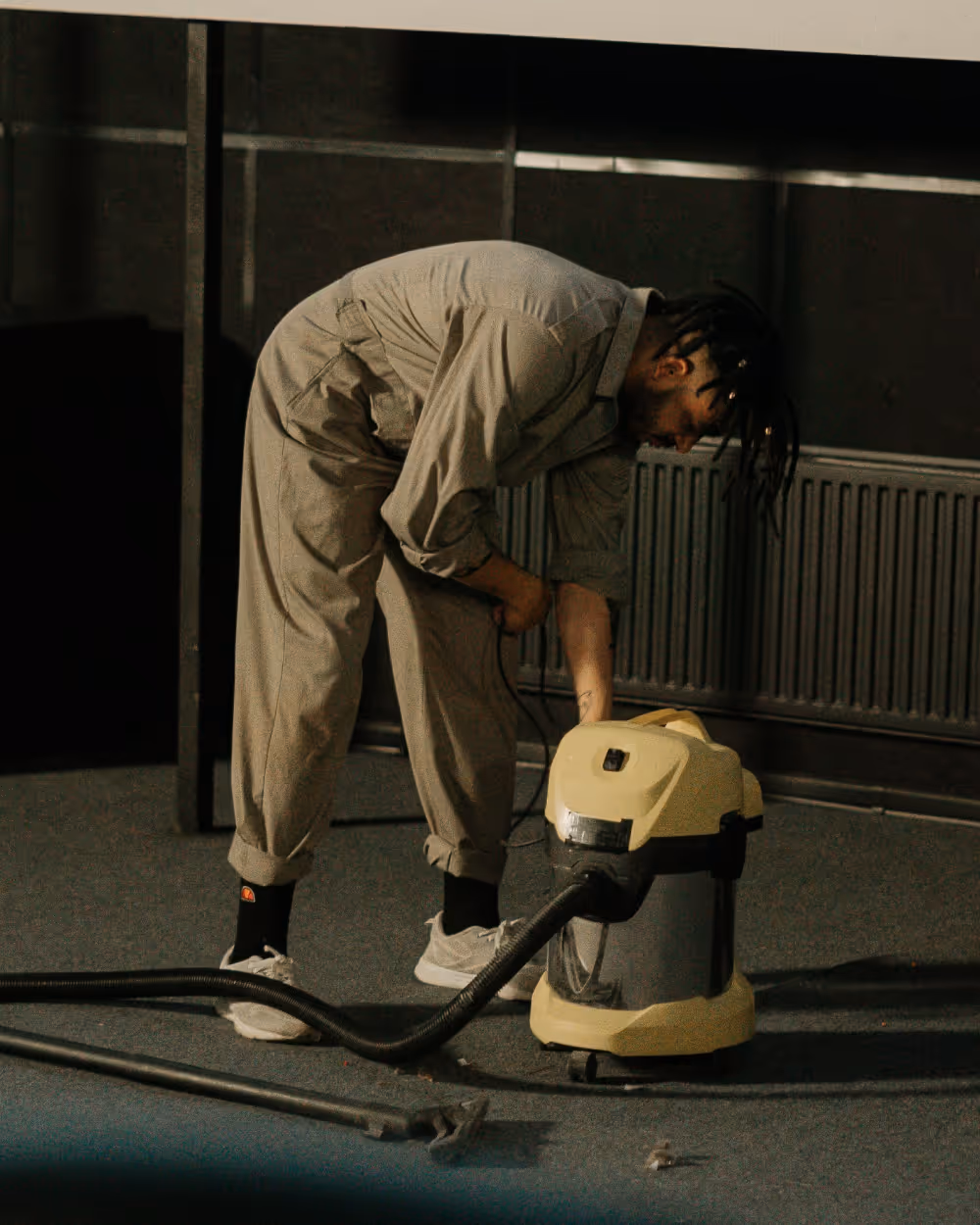 Person in beige coveralls bending over and plugging in a yellow canister vacuum cleaner on a carpeted floor.