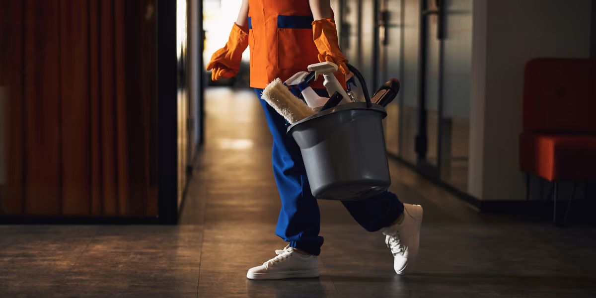 Person in orange gloves and apron carrying a bucket with cleaning supplies walking down a hallway.